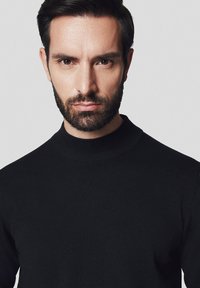 Czarny sweter z dzianiny z wysokim okrągłym dekoltem. Gładka tekstura i dopasowany krój. Model ma ciemne włosy i brodę, na neutralnym tle.