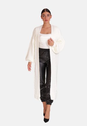 Lang, åben cardigan i hvid strik, med chunky struktur og drop-ærmer, parret med sorte plisserede joggers og spidse hæle.
