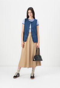 Chaleco de mezclilla con botones, camiseta de rayas azul y blanca, falda beige, bolso negro y zapatillas negras con cordones crema, de pie sobre un fondo blanco.