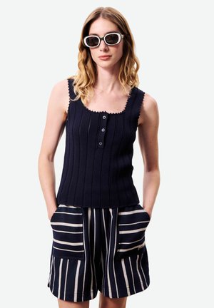 Femme aux cheveux blonds ondulés portant de grandes lunettes de soleil à monture blanche, un haut sans manches noir avec boutons, et un short bleu marine à rayures blanches verticales avec poches.