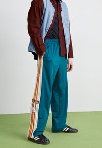 Teal träningsbyxor med vita och orange ränder på sidan, featuring tryckknappar och en Adidas logotyp patch. Matchas med en jeansväst.