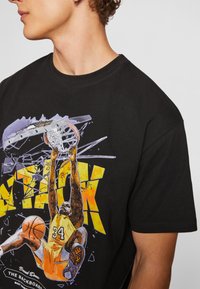 Čierne bavlnené tričko s farebnou grafikou basketbalistu, ktorý dunkuje, s nápisom "ÚTOK" v tučnom žltom a oranžovom písmene.
