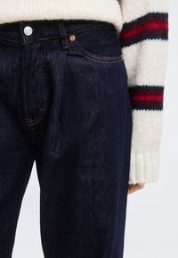 Κοντινή λήψη σκούρου μπλε τζιν με πορτοκαλί ραφές, φορεμένα με εκρού πουλόβερ που έχει κόκκινες και ναυτικές ρίγες στο μανίκι.