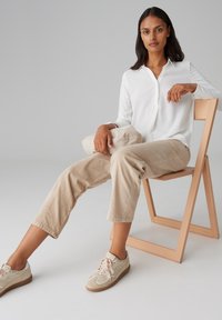 Witte lange mouwen shirt, beige corduroy broek, neutrale sneakers met lichtroze veters, zittend op een houten stoel met een beige tas in de hand.