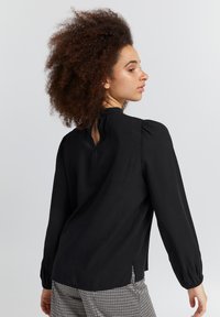 Blusa nera a maniche lunghe con collo alto, maniche a sbuffo e dettaglio a chiave sul retro. Tessuto liscio con una vestibilità comoda.