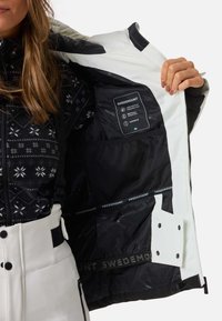 Giacca esterna bianca con fodera interna nera, dotata di un top in pile decorato con motivi di fiocchi di neve, tasche con zip e etichette per impermeabilità e traspirabilità.