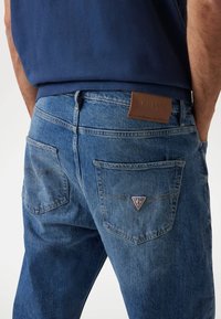 Blue denim jeans feature a leather patch with "GUESS" and a triangular logo on the back pockets, displaying a faded texture and design.