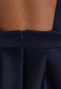 Dettagli di un abito da marinaio realizzato in un tessuto liscio, con chiusura a zip sul retro e design a taglio che mette in risalto la texture della pelle sottostante.
