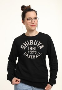 Černá mikina z měkké tkaniny s kulatým výstřihem a dlouhými rukávy, na kterých je tištěný nápis "SHIBUYA 1961 TOKYO BASEBALL" ve světle šedé barvě.