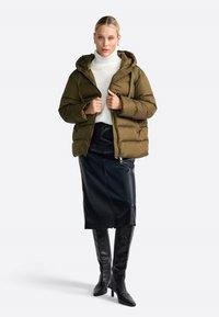 Femme portant une doudoune olive, un pull à col roulé blanc, une jupe en cuir noir et des bottes noires jusqu'au genou, se tenant devant un fond blanc.
