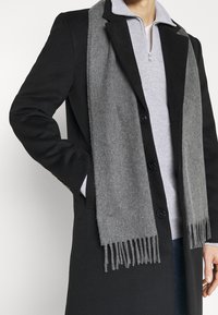 Schwarzer Mantel mit einem grauen Schal. Der Schal hat fransige Kanten, während der Mantel ein strukturiertes Design und eine durch Knöpfe geschlossene Vorderseite aufweist.