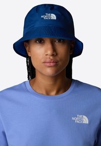 Blå buckethatt med en strukturerad yta, med en vit logotyp från The North Face framtill. Buren med en ljusblå t-shirt med rundhals.