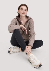 Jonge vrouw, zittend met gekruiste benen, draagt een beige fleece jack, zwarte leggings en witte sportschoenen tegen een effen achtergrond.
