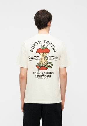 Άτομο που φοράει ένα λευκό μπλουζάκι με γραφικό ενός μήλου με έναν φίδι τυλιγμένο γύρω και το κείμενο "Earth Trippin," "Volcom Stone," και "Temptations and Libations."