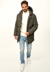 Dunkelgrüne gepolsterte Jacke mit Kapuze, Reißverschluss und Seitentaschen, getragen über einem weißen Zopfmuster-Pullover und hellblauen Jeans. Weiße Sneaker.