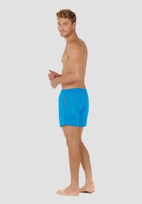 Modré plavkové šortky vyrobené z lehké tkaniny, s jednoduchým designem, elastickým pasem a bočními kapsami. Model stojí naboso a otočený do strany.
