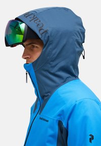 Blaue Skijacke mit dunkelblauer Kapuze, Reißverschluss und Peak Performance Logo. Glatte Textur, für Outdoor-Aktivitäten konzipiert.