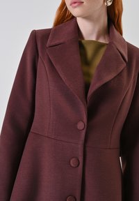 Cappotto borgogna con colletto a punte, tessuto strutturato, tre bottoni e silhouette svasata, abbinato a una maglietta verde sotto.