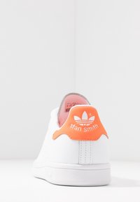 White leather sneaker with orange heel tab featuring "stan smith" logo and gray inner lining. Smooth texture and minimalist design.