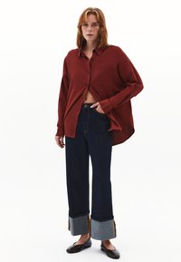 Camicia rossa con bottoni, dotata di taschino sul petto, abbinata a pantaloni in denim scuro con risvolti blu a contrasto e design a gamba dritta.