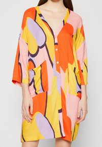Kolorowa bawełniana sukienka z dekoltem w serek, rękawami o długości trzy czwarte, dwiema przednimi kieszeniami oraz śmiałym abstrakcyjnym wzorem w odcieniach pomarańczowego, różowego i żółtego.