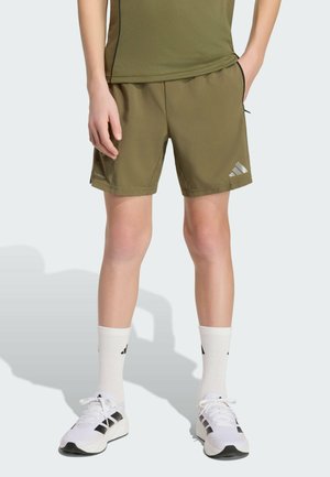 Personne portant un short de sport vert olive, des chaussettes blanches et des baskets blanches, debout avec une main dans la poche sur un fond clair.