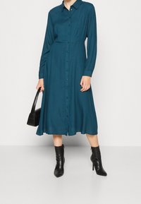 Tyrkysové dlouhé rukávové košilové šaty s obepnutým korzetem a rozšířenou sukní, spárované s černými vysokými botami na podpatku a texturovanou kabelkou.