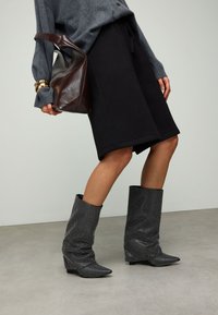 Bottes noires montantes jusqu'au genou avec un design décontracté et des pois blancs, bouts pointus, portées avec un pull gris et un short noir. Sac marron inclus.