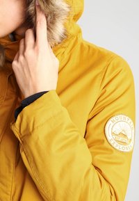 Žlutá izolovaná bunda s kapucí lemovanou kožešinou, která má na rukávu kulatou nášivku s logem a hladký, matný povrch.