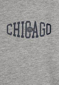 Pelēks džemperis ar "CHICAGO" uzrakstu tumši jūras zilā krāsā, iekļaujot dimanta formu un "1992" iekšpusē, uz teksturēta kokvilnas materiāla.