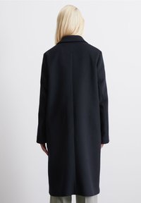 Abrigo de lana negro con corte recto, cuello muesca y mangas largas. La parte posterior cuenta con una costura que corre verticalmente por el centro.