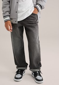 Persona que lleva puesta unos vaqueros desgastados en tonos gris y negro, zapatillas blancas y negras, una camisa blanca y una chaqueta de universidad gris con puños a rayas.
