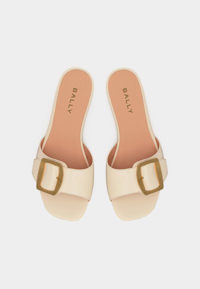 Mules beige à bouts ouverts avec larges bandes et grandes boucles dorées, semelles intérieures couleur fauve marquées du logo "BALLY", disposées côte à côte sur fond blanc.