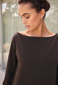 Maglia marrone a maniche lunghe con ampio scollo a barca, caratterizzata da una texture liscia e un design minimalista. Nessun motivo o dettaglio metallico visibile.