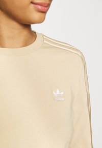 Beige långärmad sweatshirt med ribbat halsringning, kontrasterande texturerade ränder på axlarna och vit broderad logotyp på övre vänstra bröstet.