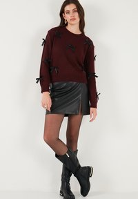 Burgunderfarbener Pullover mit schwarzen Bandakzenten, runder Ausschnitt; kombiniert mit einem schwarzen Leder-Minirock mit seitlichem Schlitz und kniehohen schwarzen Stiefeln.