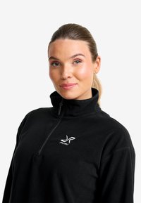 Svart fleece-pullover med hög krage och halvmångdragare, som har en vit logotyp på vänster bröst och en mjuk, texturerad yta.