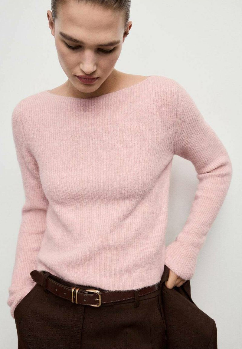 Donna che indossa un morbido maglione rosa a costine infilato in pantaloni marrone scuro con una cintura abbinata, che guarda in basso con le mani nelle tasche.