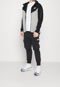 Hombre con sudadera con cremallera de Nike en negro y gris, camiseta blanca, pantalones cargo negros y zapatillas blancas y grises, de pie sobre un suelo gris.
