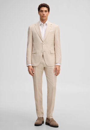 Giovane uomo in piedi che indossa un completo beige con camicia bianca e scarpe in camoscio marroni su uno sfondo chiaro uniforme.