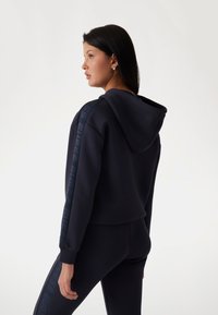 Sweat à capuche cropped bleu marine avec une capuche, des poignets côtelés et un logo Guess le long des manches. Fabriqué en tissu doux et lisse avec une coupe ajustée.