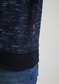 Sudadera azul marino con un patrón texturizado, que presenta líneas blancas contrastantes y una etiqueta naranja, terminada con un dobladillo acanalado en azul marino.