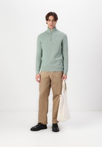 Light green, ribbed knit sweater with half-zip collar, paired with tan trousers and black shoes. White tote bag held in left hand.