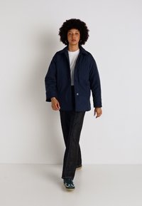 Giacca blu navy con chiusura a bottoni, grandi tasche e vestibilità rilassata, abbinata a pantaloni neri texturizzati e sneakers verdi.