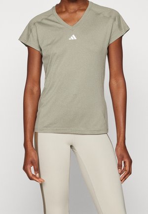 Kobieta ubrana w jasnooliwkową, krótki rękaw, sportową koszulkę z dekoltem w serek i białym logo Adidas oraz beżowe dopasowane legginsy sportowe.