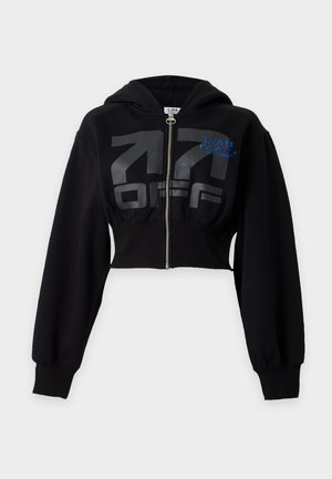 Femme aux longs cheveux bouclés porte un sweat à capuche court noir zippé, un pantalon large noir avec cordon, et des baskets noires et blanches, debout les mains dans les poches.