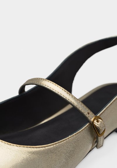 Χρυσό μεταλλικό παπούτσι slingback με μαύρη επένδυση από μαξιλαράκι και μικρή χρυσή αγκράφα στο λουράκι πάνω από το πόδι.