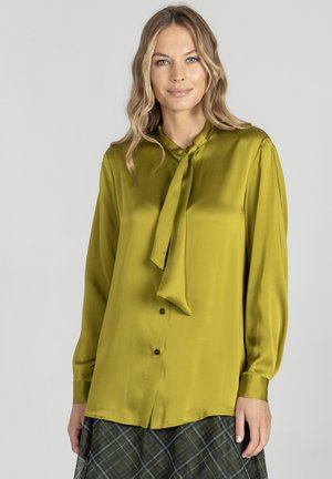 Kobieta w oliwkowo-zielonej satynowej bluzce z długim rękawem i wiązaniem przy szyi, w kratkowanej spódnicy, stojąca na tle szarego tła.