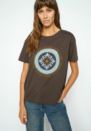 Vrouw draagt een donkerbruin t-shirt met een blauw en goud rond ontwerp en de woorden vrede, liefde, hart herhaald eromheen, gecombineerd met blauwe spijkerbroek.