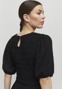 Sort kjole med tekstureret smock, puffede ærmer og en nøglehulslukning i ryggen. Blødt, krøllet stof over det hele.